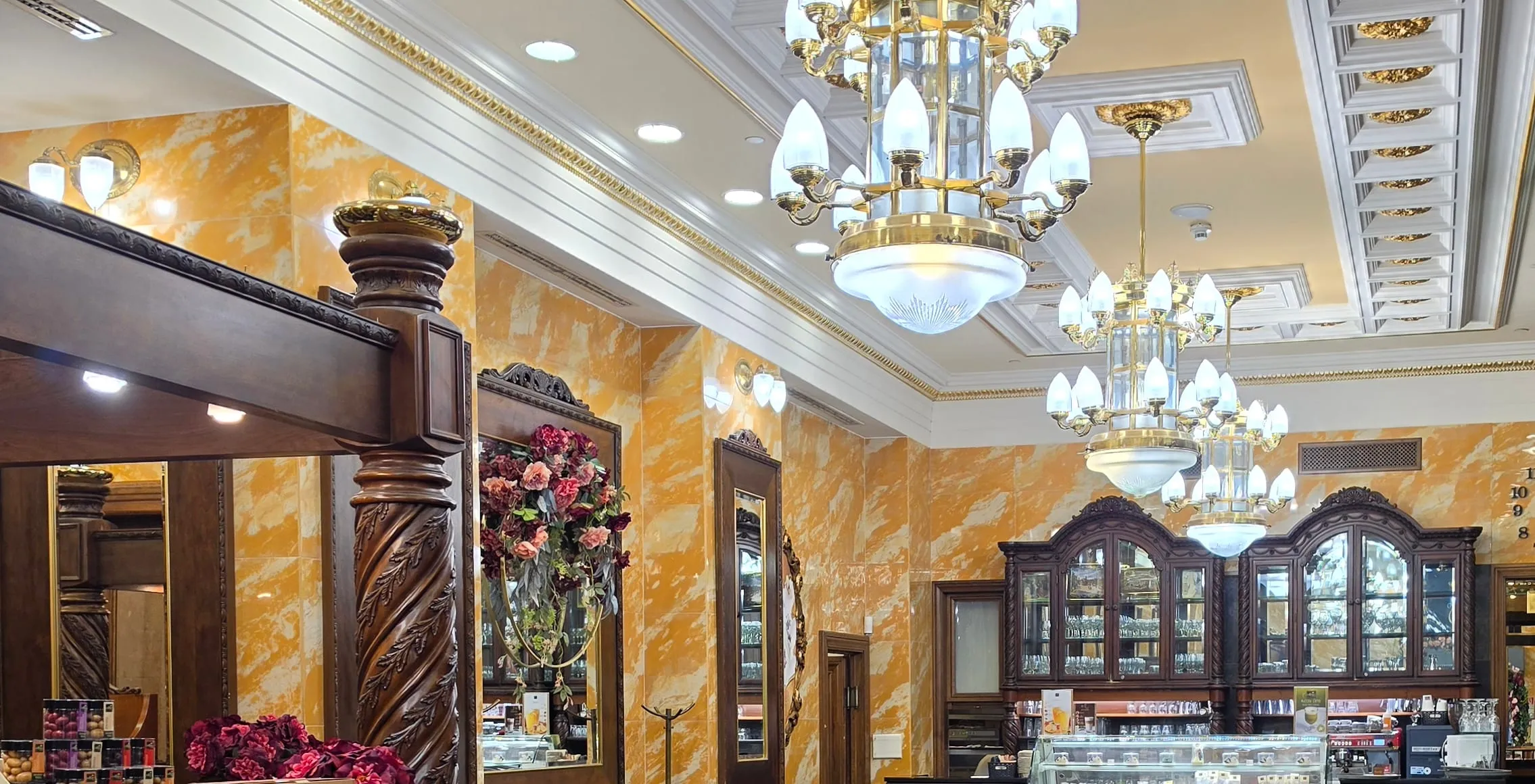 Interiér cukrárne Szamos Gourmet Ház — steny z medovožltého mramoru, mosadzné lustre s bielymi tienidlami, tmavé vyrezávané drevo, vitríny so sklom a kytica ruží