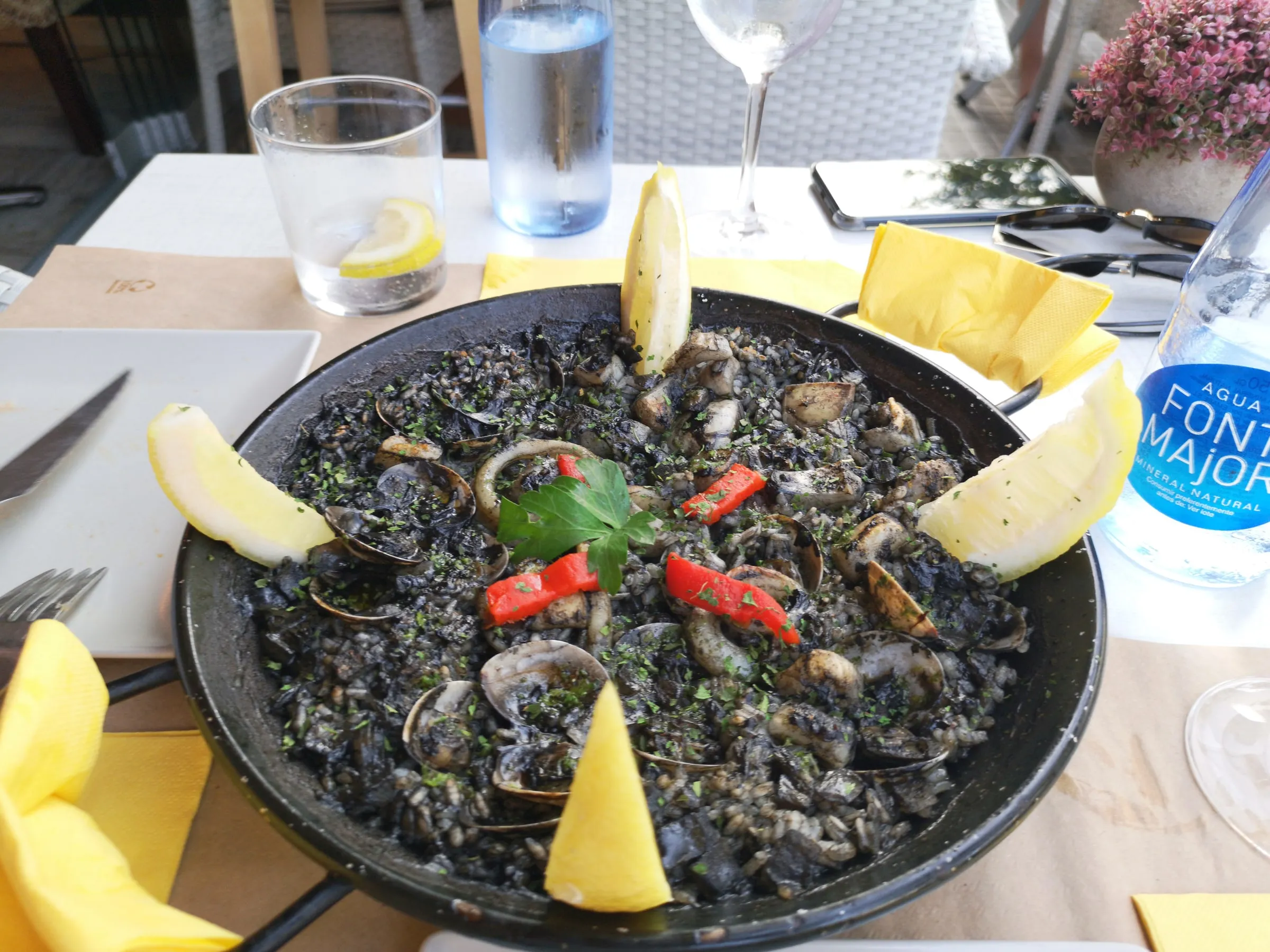 A pan of black paella, arròs negre: rice coloured with cuttlefish ink, clams, squid rings, red pepper and lemon, water and a yellow napkin on the table