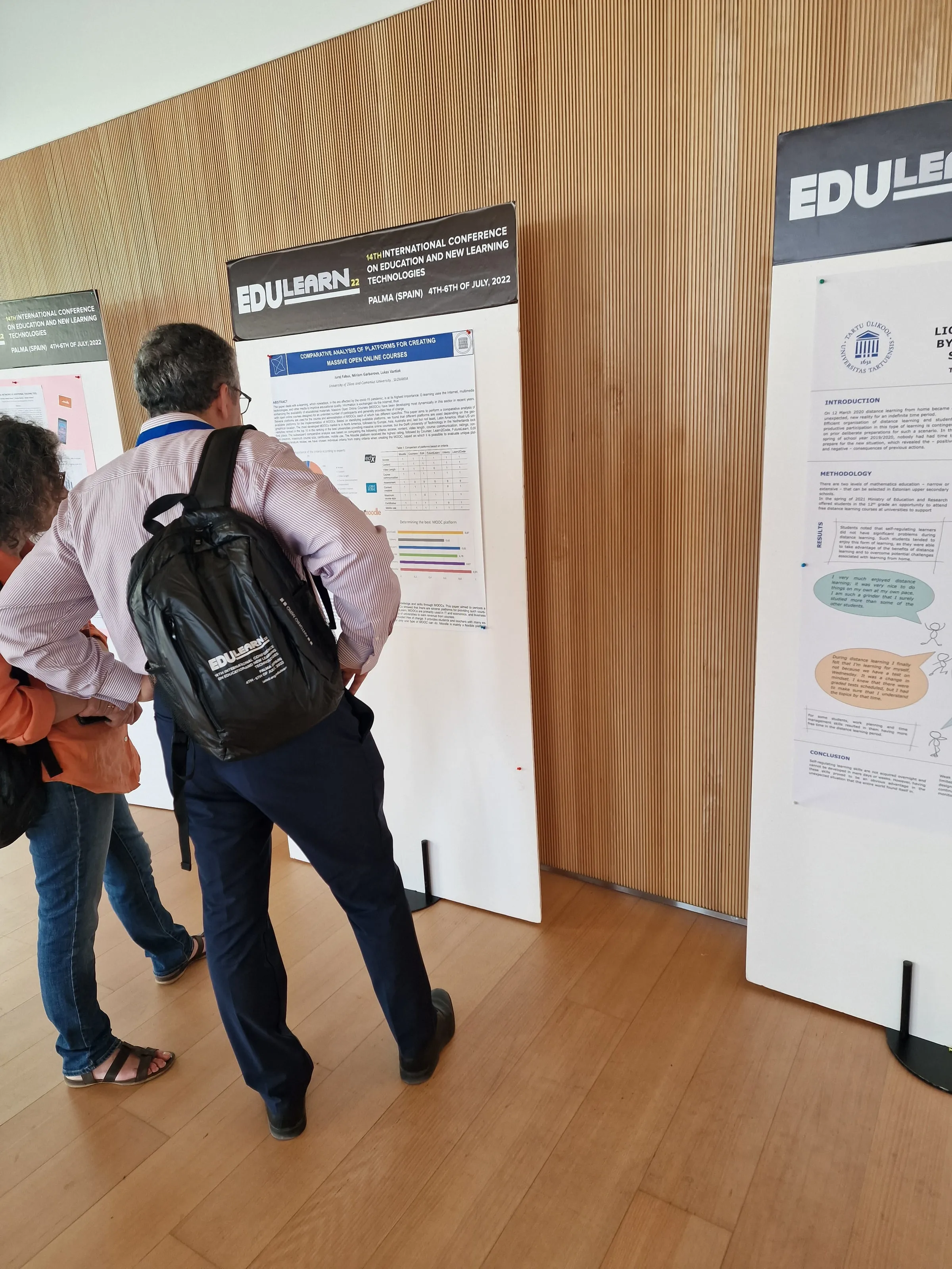 A conference participant with a backpack stands with his back to the camera reading a poster on platforms for MOOC courses at EDULEARN22; more posters on a wooden wall beside it