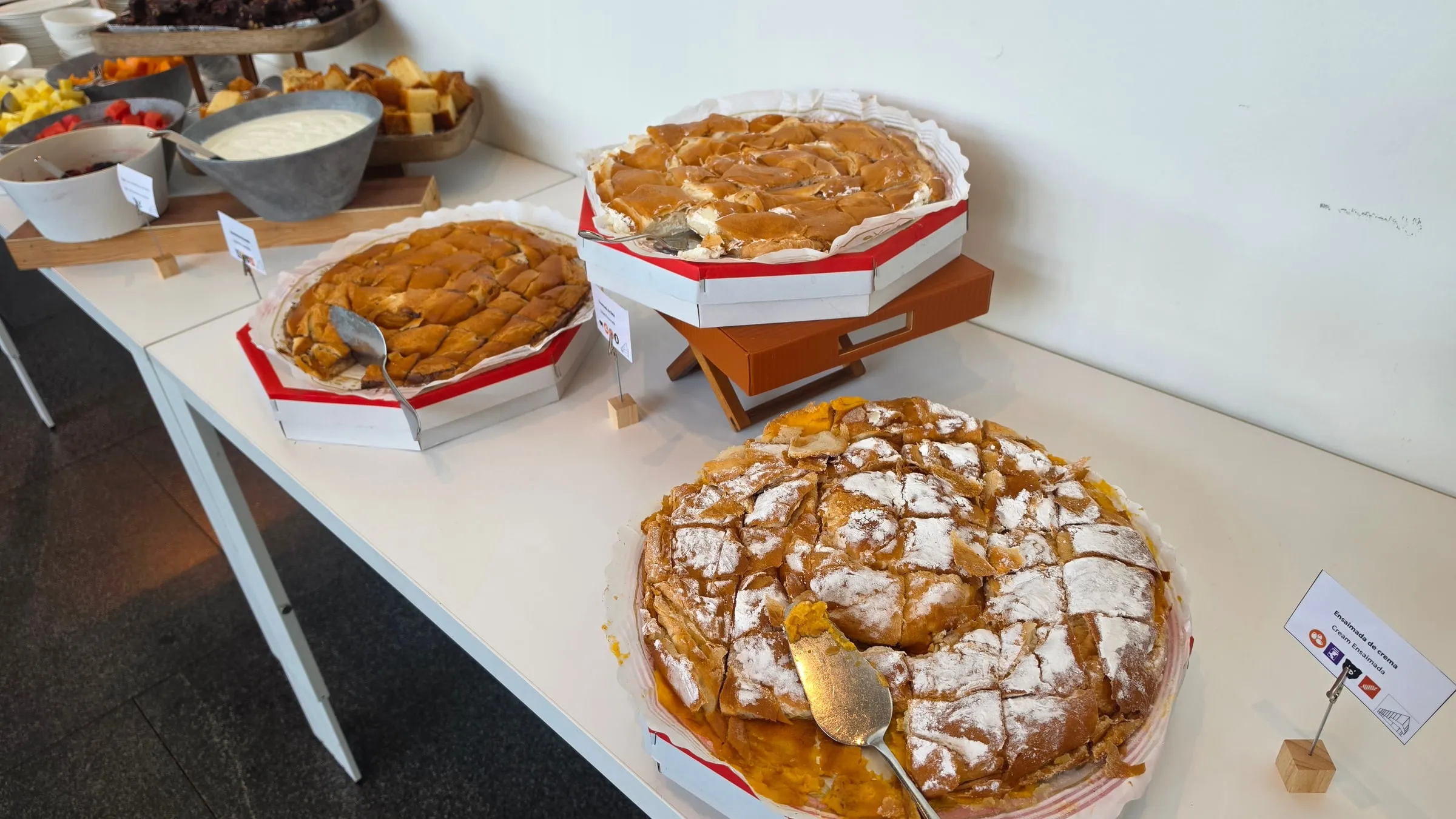 Three large ensaïmadas on the dessert table at a conference lunch — a cream ensaïmada dusted with icing sugar at the front with pieces cut out, bowls of fruit and yoghurt behind