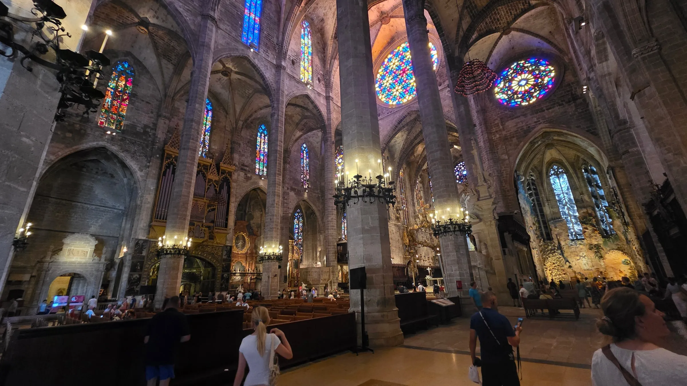 Interior of La Seu cathedral: tall stone columns, Gothic vaulting, colourful stained glass and two rose windows, pews and visitors below