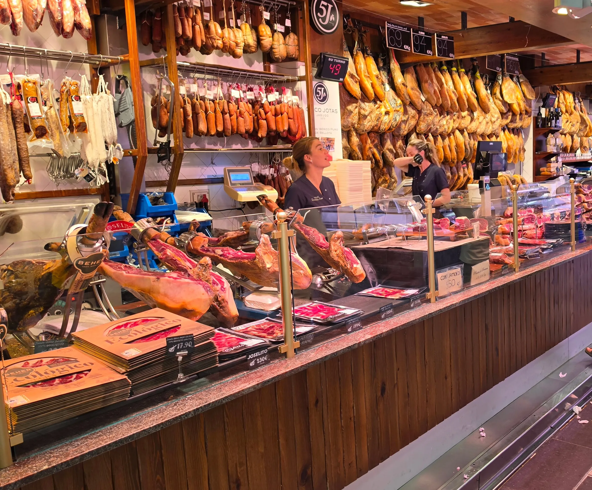 A ham stall at the Mercat de l'Olivar market: dozens of hanging jamóns and sausages, sliced cuts in the counter and shop assistants behind the glass