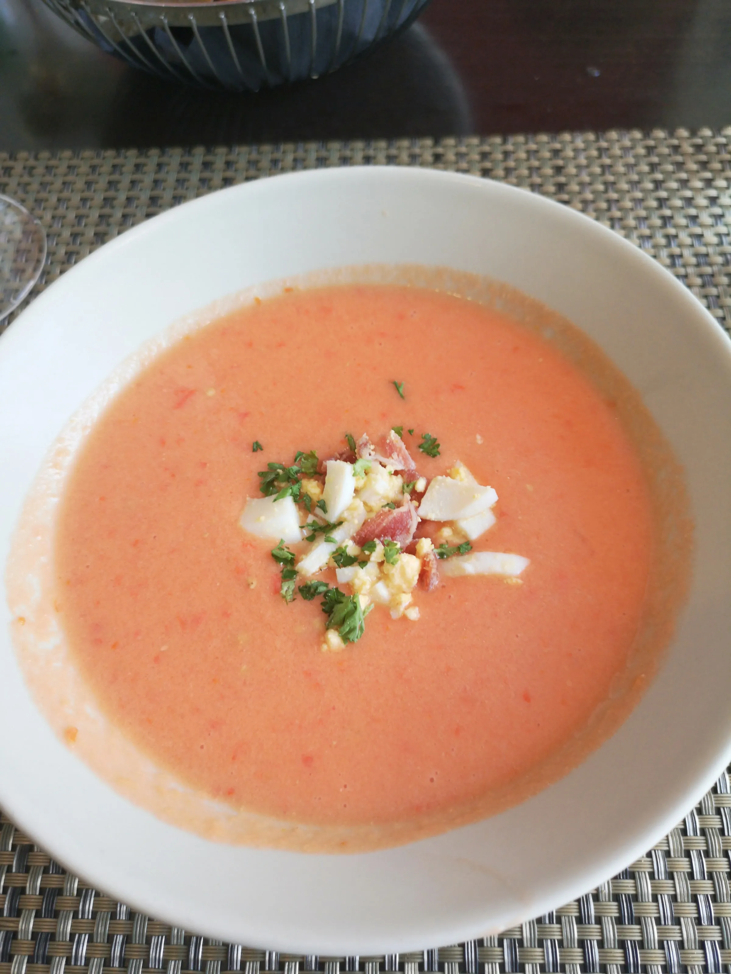 A bowl of thick orange-pink cold salmorejo soup topped with chopped egg, pieces of ham and parsley