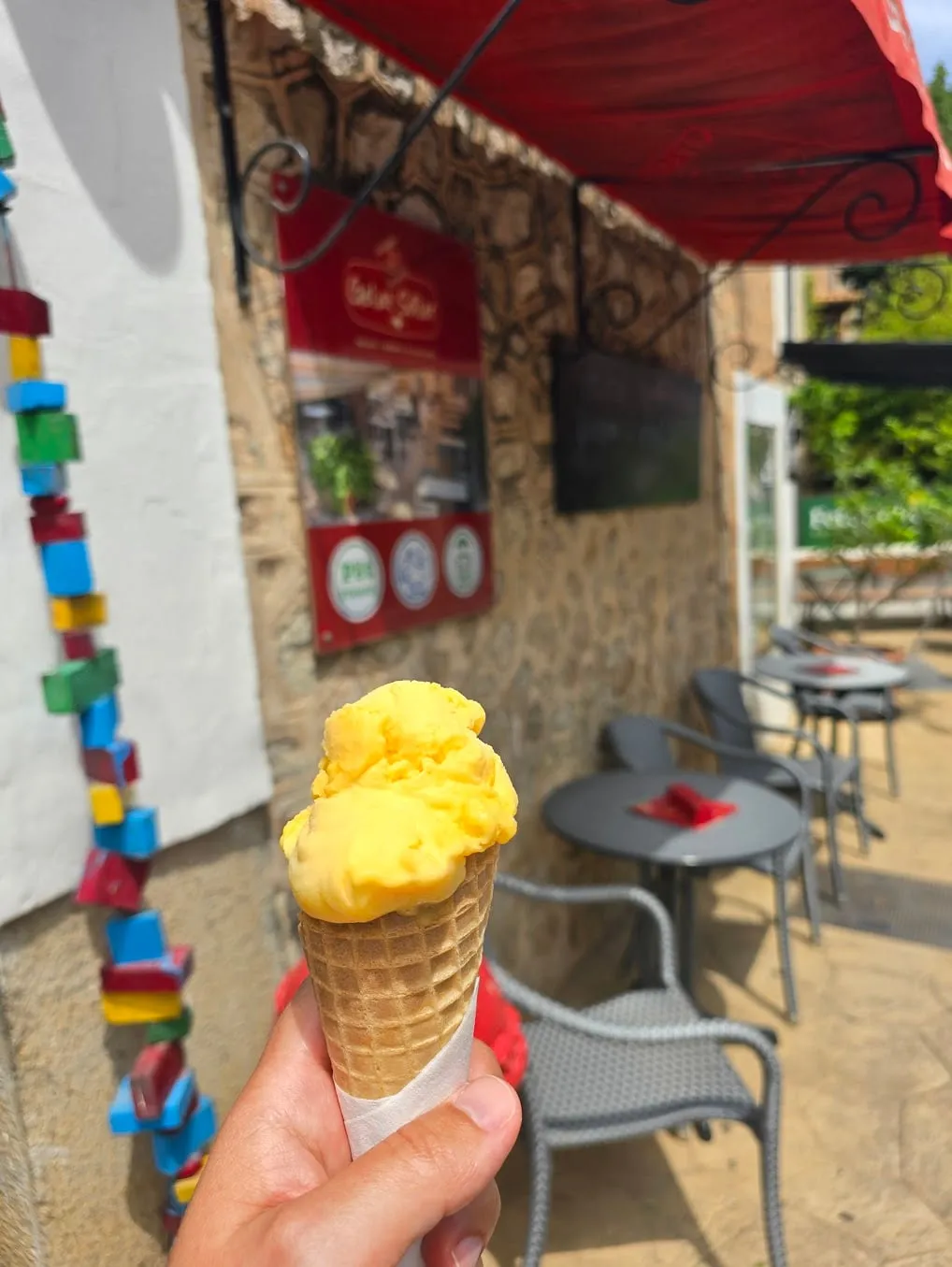 A hand holds a cone with two scoops of orange tangerine ice cream in front of the stone wall of an ice-cream shop in Sóller, tables under a red awning behind