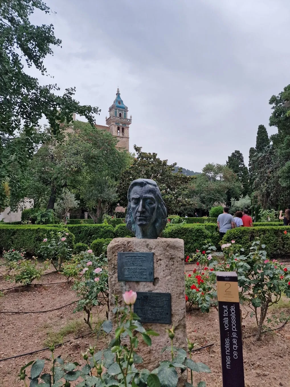 A bronze bust of Chopin on a stone plinth in the garden of the Valldemossa charterhouse, roses and clipped hedges around it, the church tower with its green-and-white ceramic roof in the background