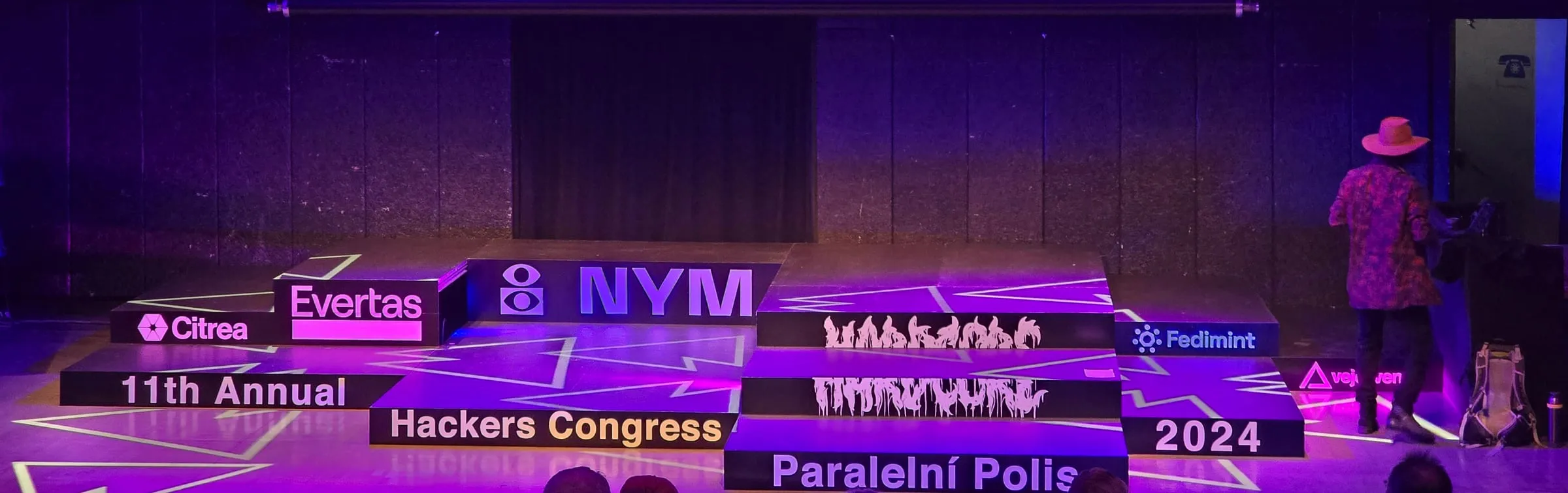Pódium kongresu HCPP vo fialovom svetle — stupňovité čierne kvádre s nápismi 11th Annual, Hackers Congress, Paralelní Polis a 2024 a logami partnerov, na podlahe svietiace línie, vpravo muž v ružovom klobúku otočený chrbtom