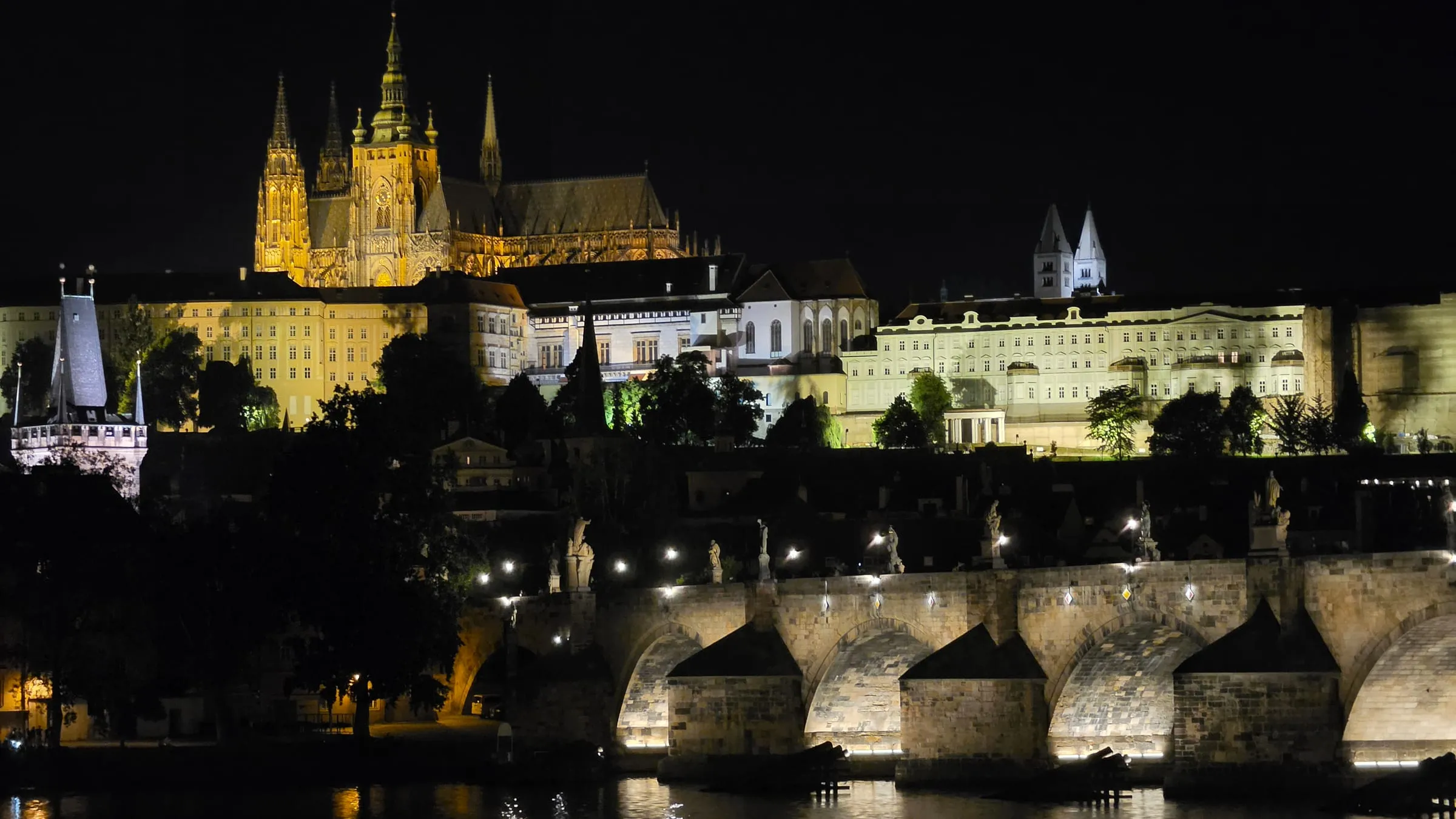 Nočný pohľad cez Vltavu na osvetlený Pražský hrad — hore Katedrála svätého Víta v zlatom svetle, pod ňou dlhé fasády hradných palácov a dve biele veže Baziliky svätého Juraja, vpravo dole Karlov most s lampami a sochami, v rieke odlesky svetiel