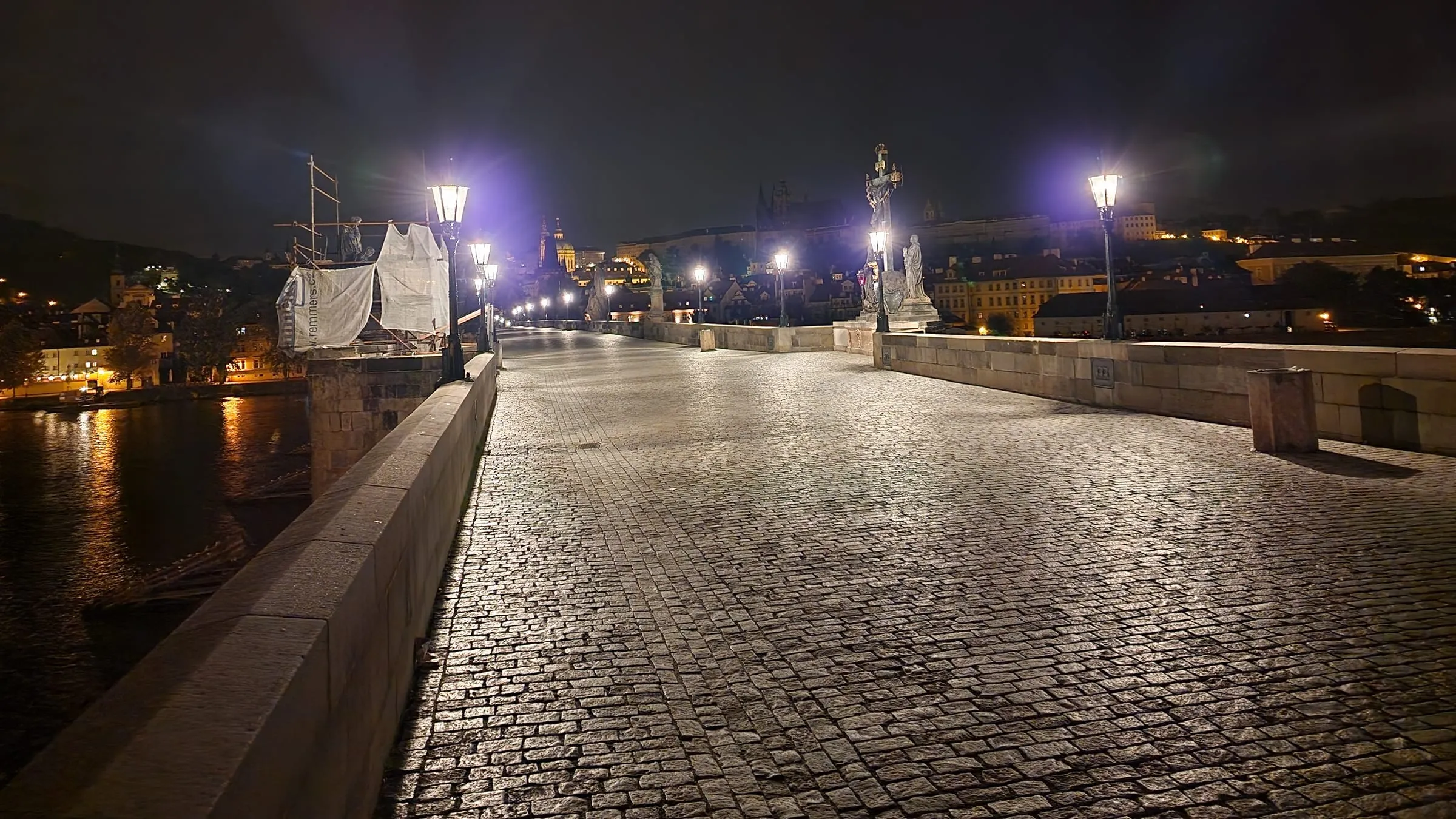 Prázdny Karlov most v noci — lesklá dlažba z mačacích hláv vedie k Malej Strane, lampy s fialovou žiarou, barokové sochy na zábradlí, vľavo lešenie zakryté plachtou a Vltava s odleskami svetiel, v pozadí tmavá silueta Hradu