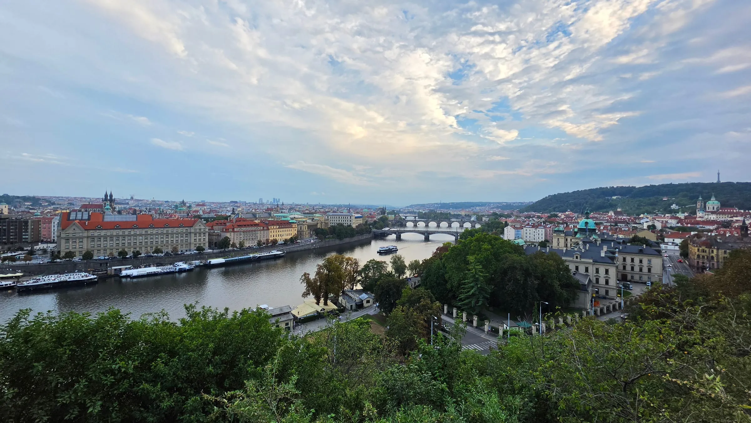 Podvečerný pohľad z Letnej na Vltavu, ktorá sa stáča do diaľky pod radom mostov, na brehoch nábrežia s loďami a historická zástavba, vzadu zalesnený Petřín s rozhľadňou, nad tým dramatická obloha s roztrhanými oblakmi