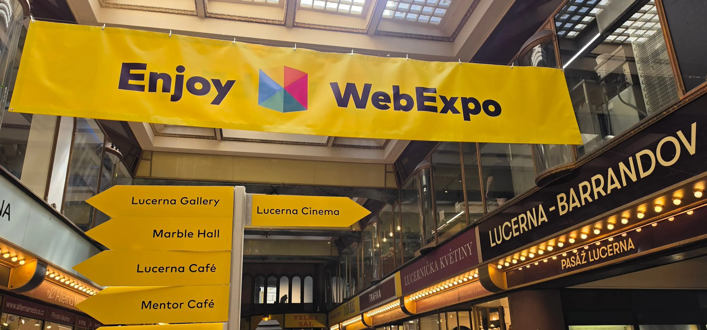 Pasáž Lucerna počas Webexpa — pod presklenou strechou žltý transparent Enjoy WebExpo, pod ním žlté smerové šípky Lucerna Gallery, Marble Hall, Lucerna Café, Mentor Café a Lucerna Cinema, vpravo svietiace nápisy Lucerna-Barrandov a Pasáž Lucerna