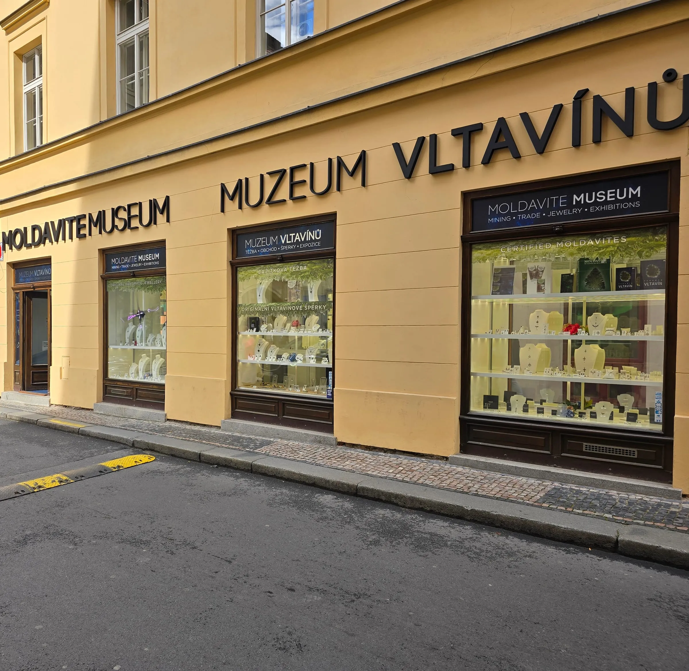 Žltá fasáda s výkladmi a veľkým čiernym nápisom Muzeum vltavínů a Moldavite Museum, vo výkladoch šperky s vltavínmi na bielych stojanoch, pred budovou dláždený chodník a ulica so žlto-čiernym spomaľovačom