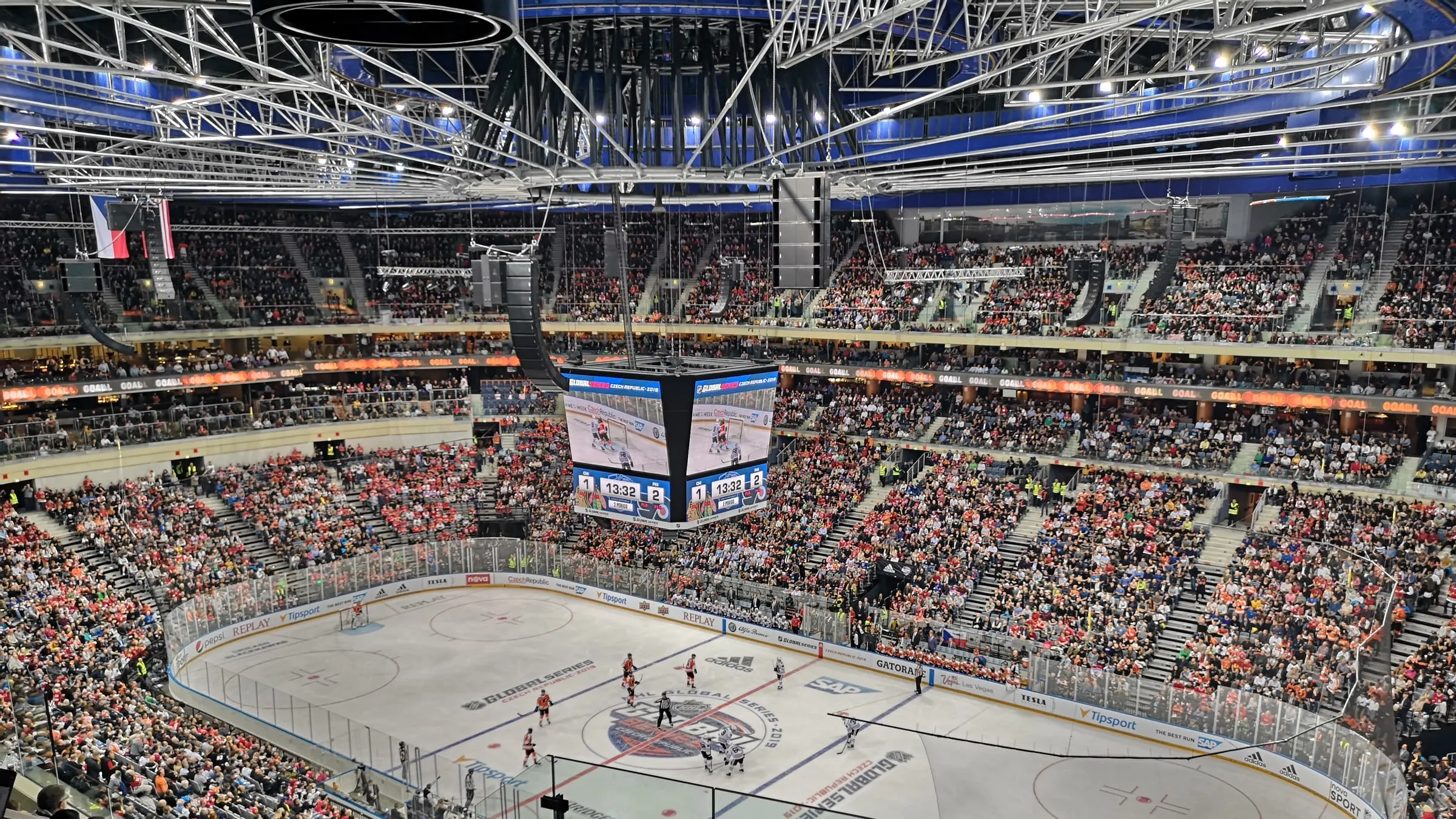 Pohľad z horného radu do zaplnenej O2 areny počas zápasu NHL Global Series — biela ľadová plocha s logami Global Series, hráči v oranžových a bielych dresoch, nad stredom kocka s obrazovkami, tribúny plné fanúšikov v oranžovom a červenom, hore oceľová strešná konštrukcia a česká vlajka