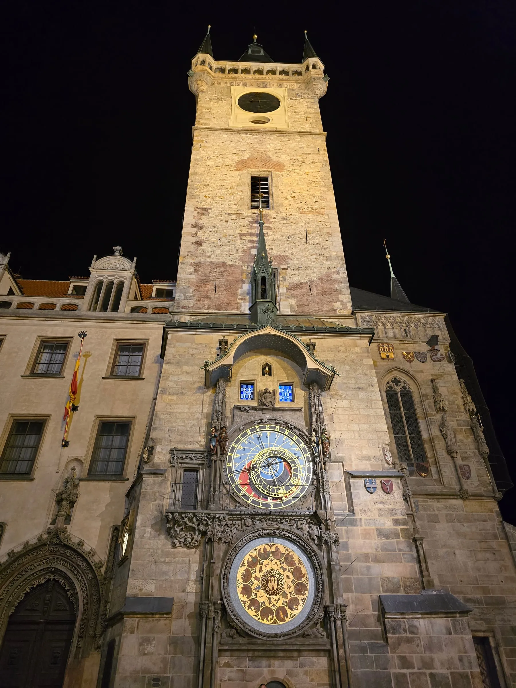 Staromestská radničná veža v noci zdola — osvetlený orloj s astronomickým ciferníkom a zlatým kalendárnym kruhom, modro svietiace okienka apoštolov, nad tým svetlá kamenná veža s hodinami, vpravo gotická kaplnka