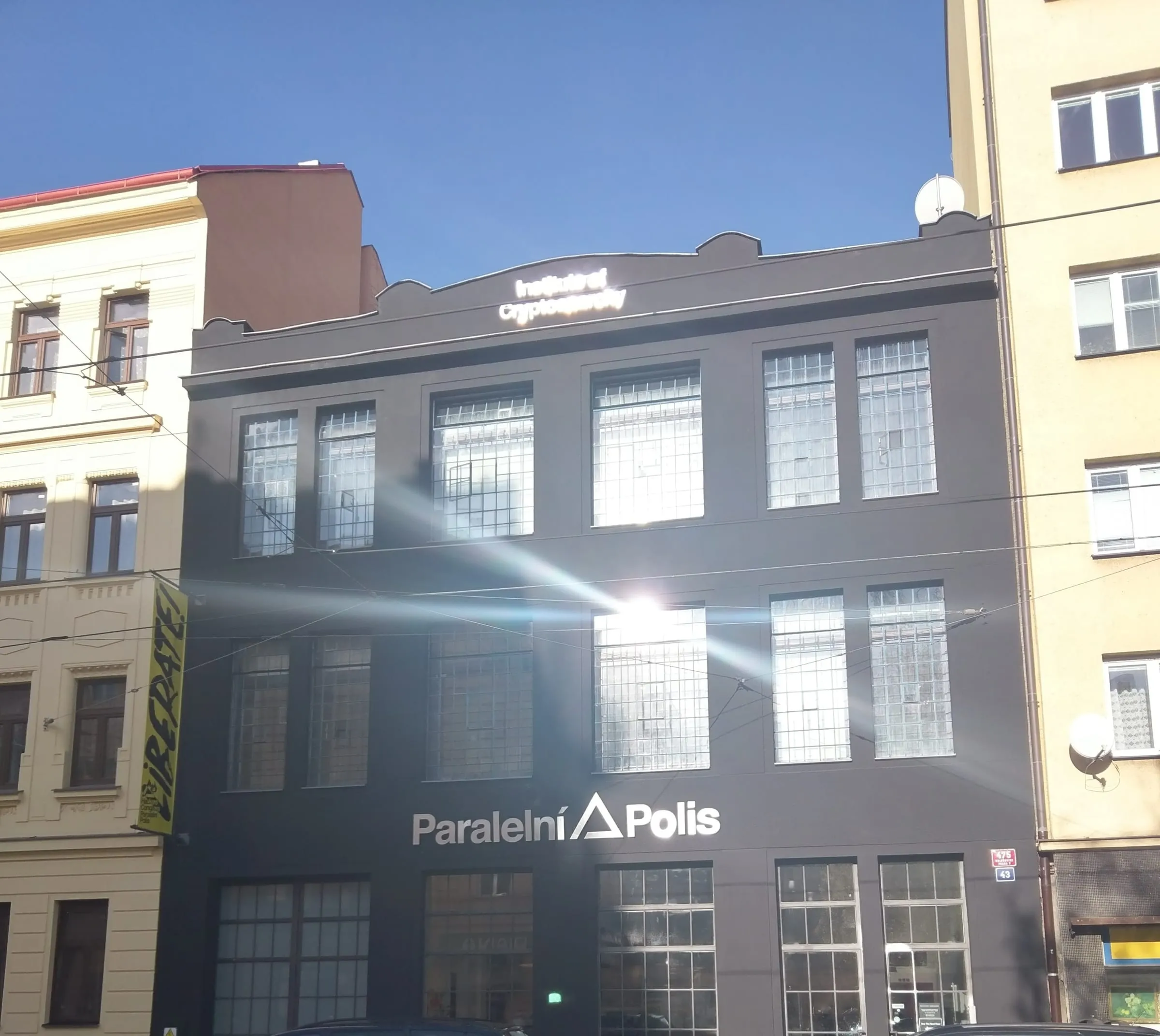 Trojpodlažná tmavosivá budova s veľkými mrežovanými oknami a bielym nápisom Paralelní Polis s logom trojuholníka na fasáde, na atike svetelný nápis Institute of Cryptoanarchy, vľavo žltý činžiak so žltým transparentom, modrá obloha a odlesk slnka