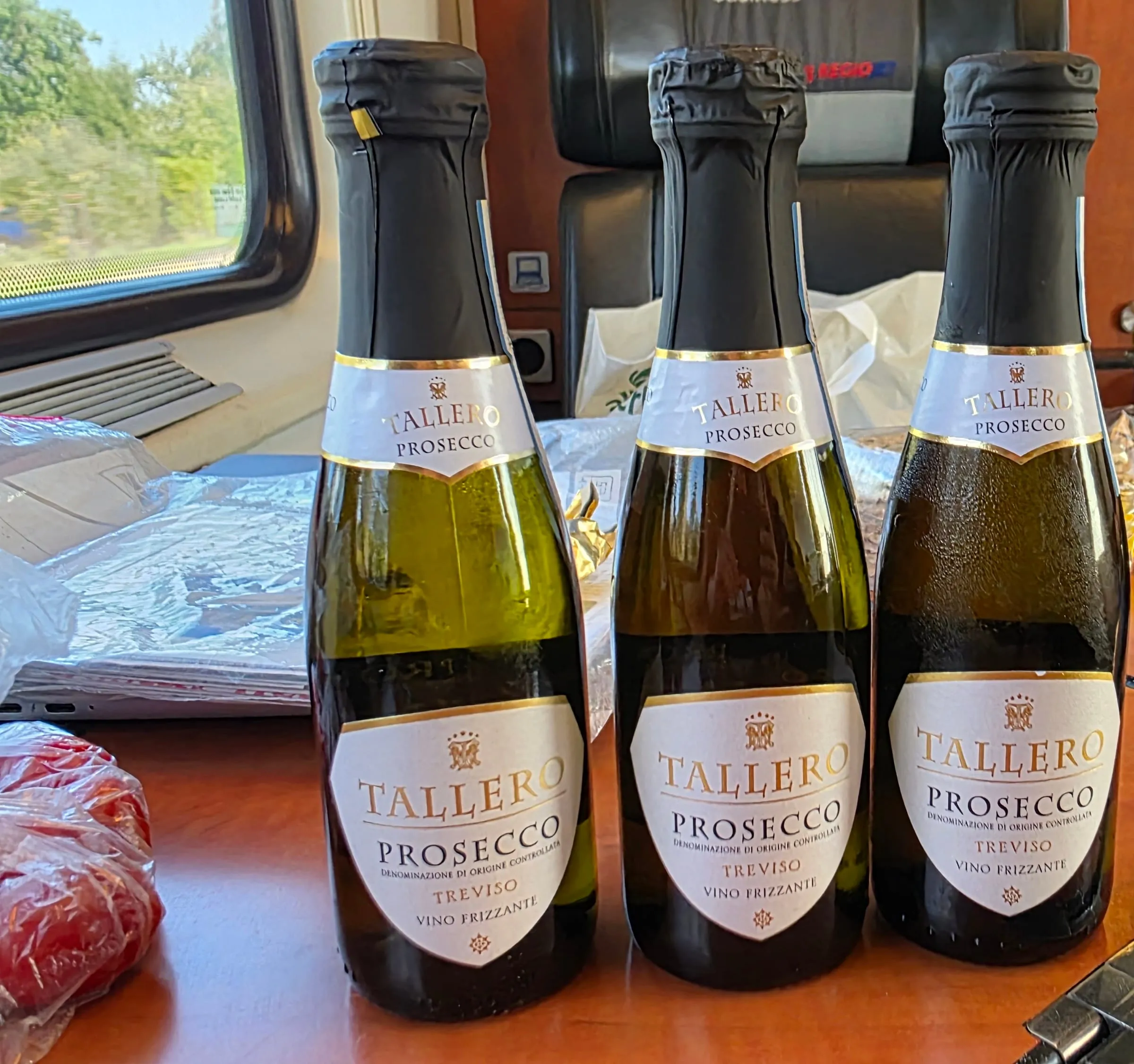 Tri malé fľaše prosecca Tallero Treviso na drevenom stolíku vo vlaku RegioJet, za nimi čierne kožené sedadlo a okno so zelenou krajinou