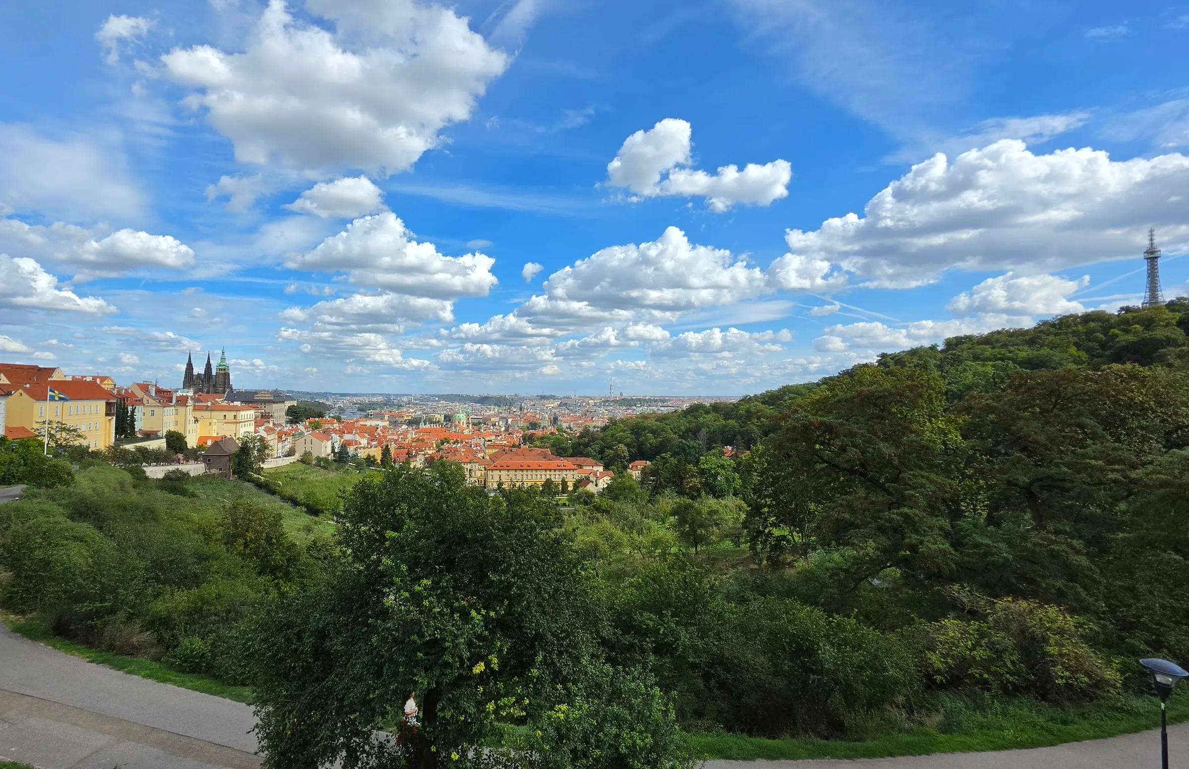 Pohľad zo svahu pri Strahovskom kláštore cez sady na Pražský hrad s vežami Katedrály svätého Víta, červené strechy Malej Strany a mesto až po obzor, vpravo zalesnený Petřín s rozhľadňou, nad tým modrá obloha s bielymi oblakmi