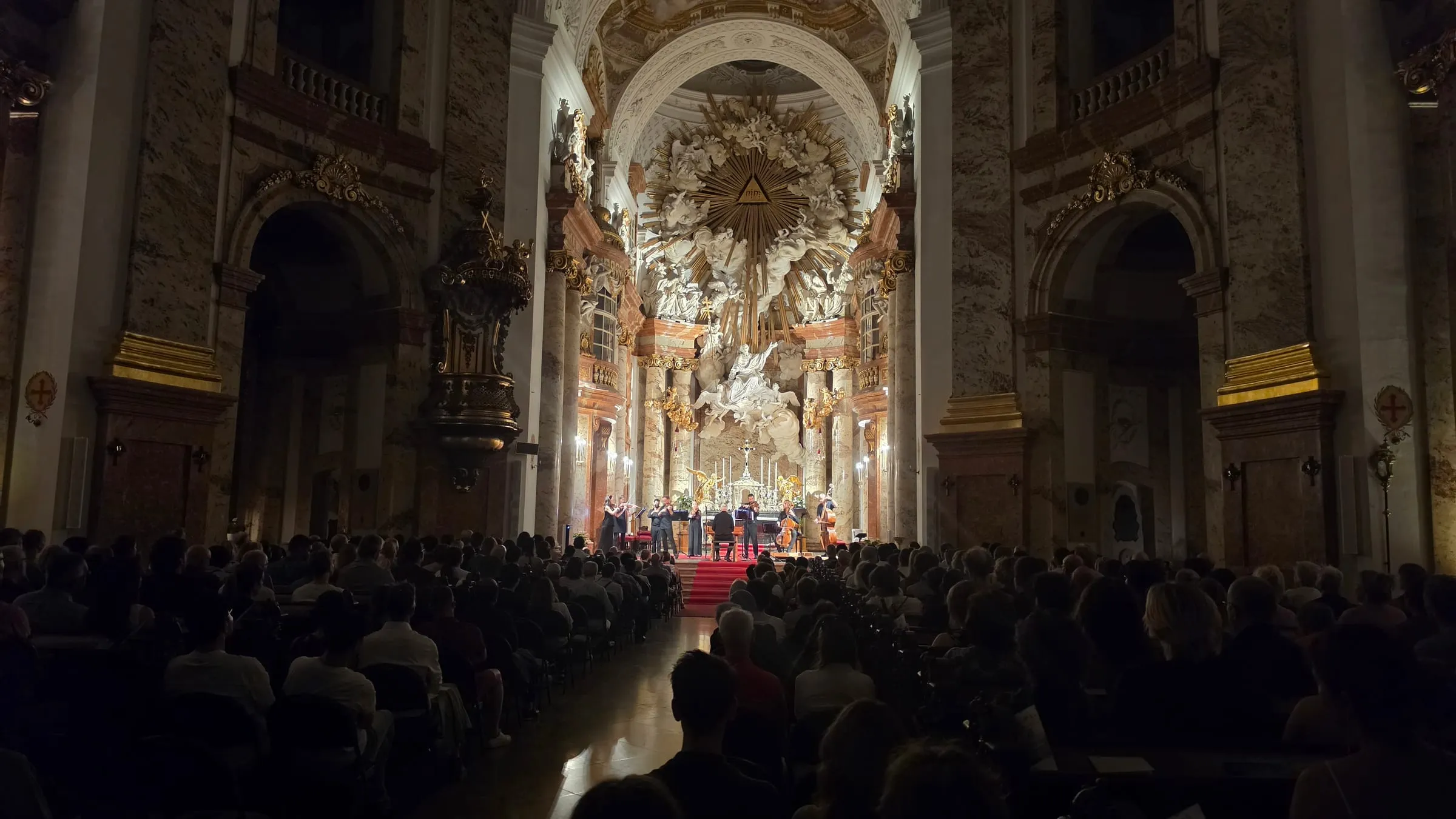 Interiér Karlskirche počas večerného koncertu — publikum odzadu, vpredu na červenom koberci malý orchester, za ním osvetlený barokový oltár so zlatým lúčovým motívom a mramorové stĺpy