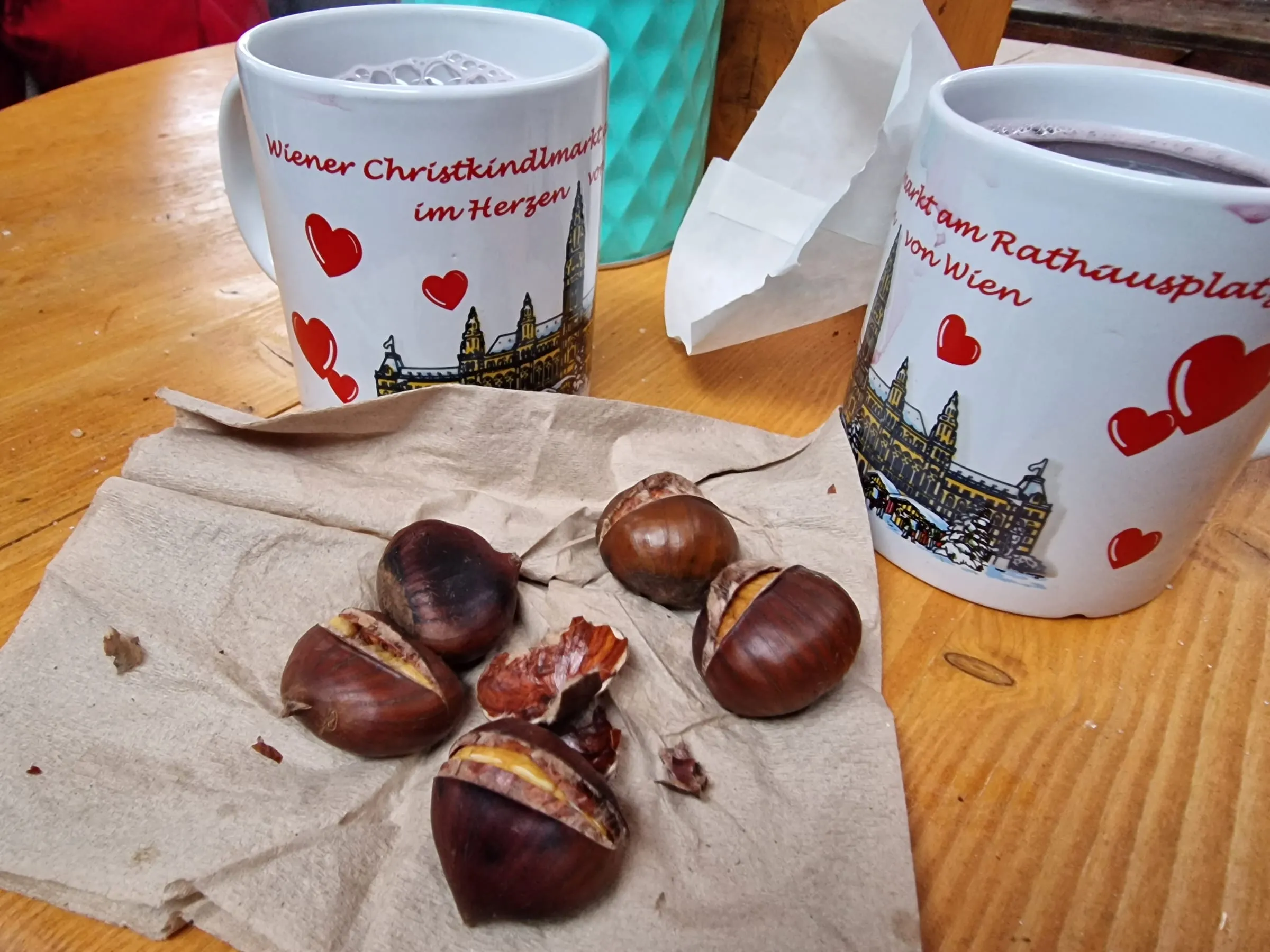 Dva biele hrnčeky s červenými srdiečkami a nápisom Wiener Christkindlmarkt na drevenom stole, pred nimi na papieri pečené gaštany