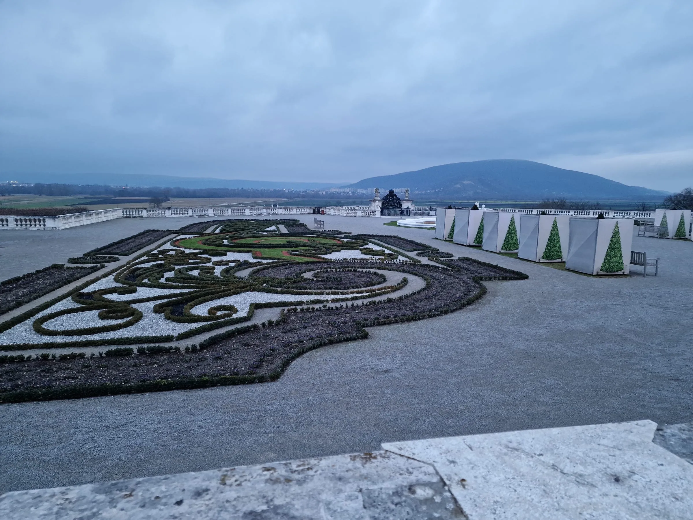 Terasová záhrada Schloss Hof v decembri — ornamentálne záhony zo zastrihnutých buxusov a štrku, vpravo stánky vianočného trhu s natlačenými stromčekmi, za balustrádou rovina Marchfeldu a kopec na obzore pod sivou oblohou