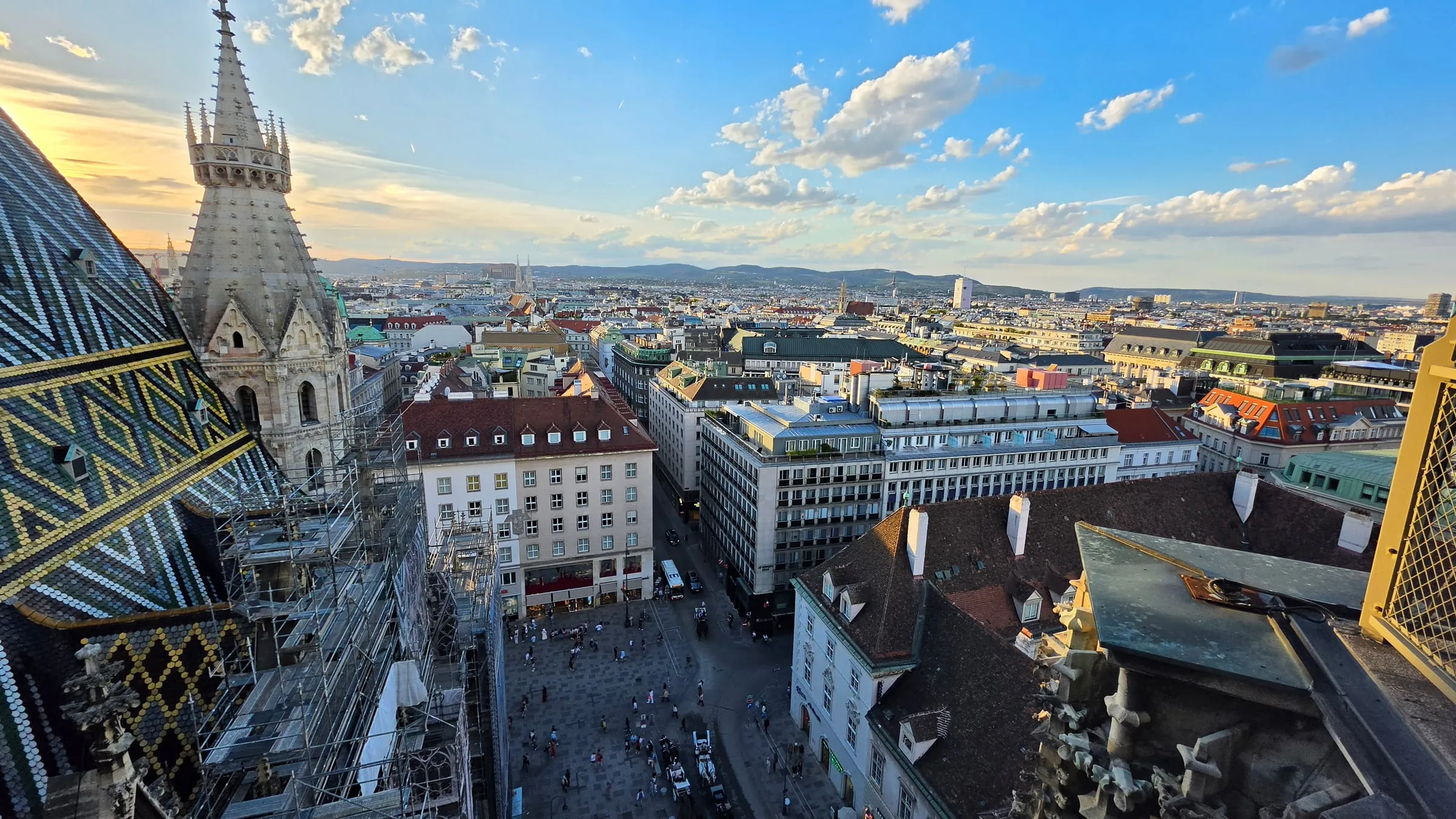 Výhľad zo severnej veže Dómu svätého Štefana — vľavo cikcaková strecha z farebných škridiel a špic južnej veže, dole Stephansplatz s ľuďmi, v pozadí strechy Viedne a kopce Viedenského lesa pri západe slnka